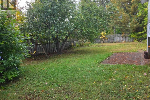 Rear view of the backyard with a concrete patio, green grass, trees, and a wooden fence. - 177 River Rd, Sault Ste  Marie