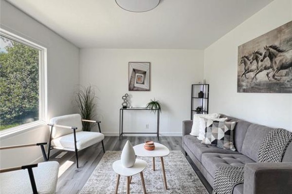 Living room with grey sofa, two white chairs, patterned rug, and a large window with natural light. - 1795 Manitoba Avenue, Winnipeg