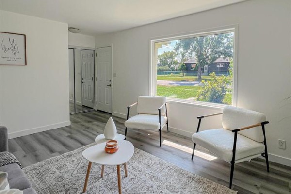 Living room with two white armchairs, a coffee table, and a large window showing a street view. - 1795 Manitoba Avenue, Winnipeg