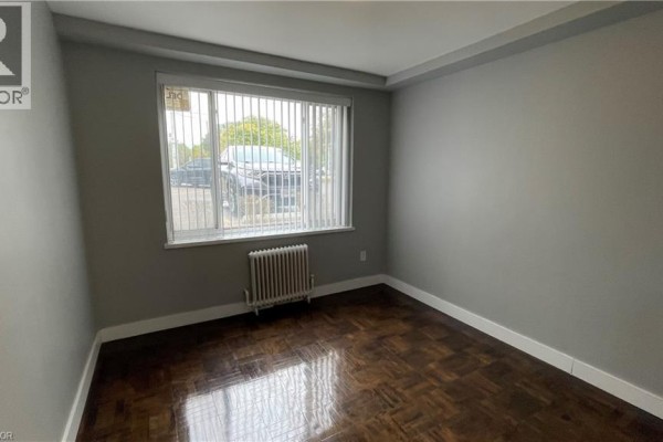 Room with dark hardwood floors, grey walls, vertical blinds, and a window overlooking the street. - 1797 Main Street W Unit# 3, Hamilton