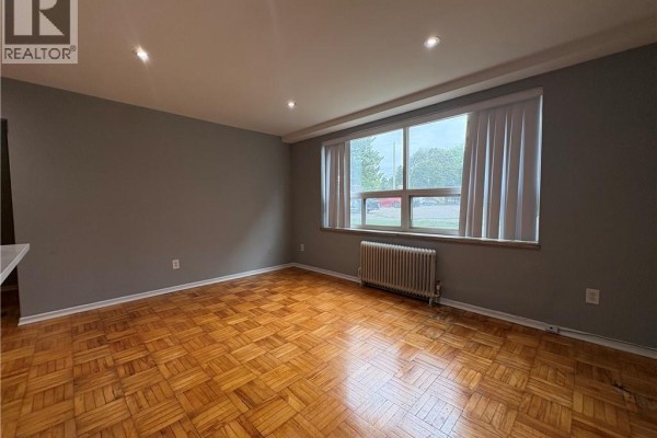 Living room with parquet flooring, grey walls, and a large window with vertical blinds and a radiator. - 1797 Main Street W Unit# 4, Hamilton