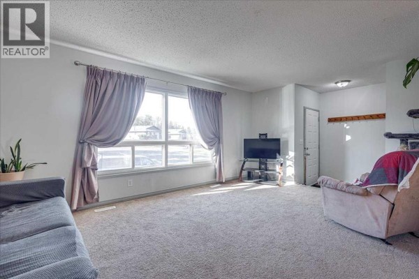 Living room featuring a large window with draped curtains, a TV stand, and a grey carpet. - 18 Greenham Drive, Red Deer
