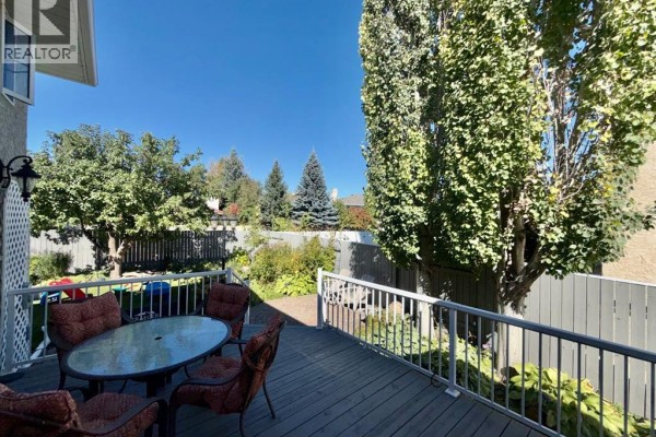 Deck with glass-topped table and upholstered chairs overlooks a fenced backyard with mature trees and shrubs. - 18 Heritage Close W, Lethbridge