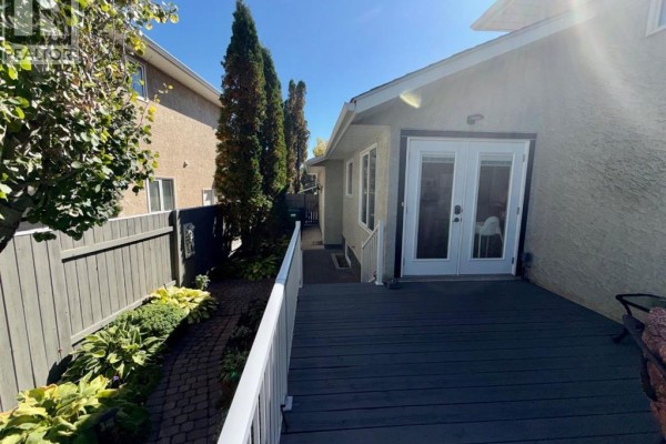 Rear exterior showing a grey wooden deck with white railings leading to double glass doors and a fenced backyard. - 18 Heritage Close W, Lethbridge