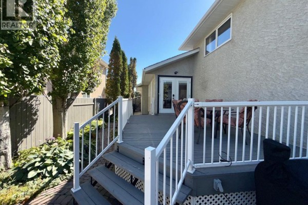 View of the backyard deck with grey composite decking, white railings, and stairs, surrounded by greenery and fencing. - 18 Heritage Close W, Lethbridge