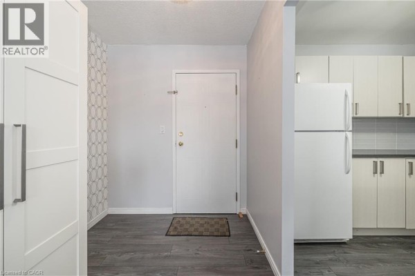 Hallway leading to apartment door with a white refrigerator and textured wallpaper. - 18 Holborn Court Unit# 211, Kitchener