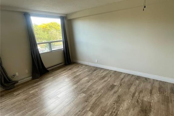 Living room with light wood laminate flooring, beige walls, and a large window with grey curtains. - 18 Holborn Court Unit# 212, Kitchener