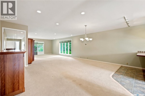 Open concept living area with neutral carpeting, large windows offering a green view, and a dining area with a built-in counter. - 18 Lakeview Place, Regina