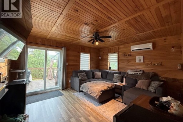 Living room with wood-paneled walls, a large sectional sofa, and a sliding glass door to the deck. - 18 Parc P'tiso Street, Edmundston