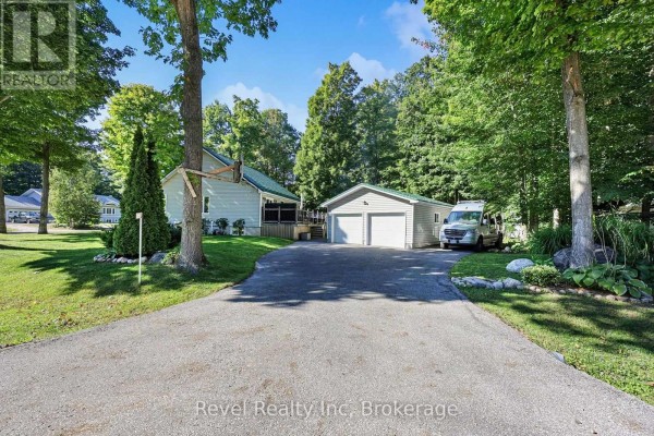 A paved driveway leads to a detached two-car garage with white doors, surrounded by trees and greenery. - 18 Rue Vanier Road, Tiny
