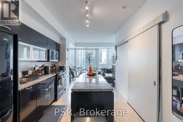 Modern kitchen with black cabinetry, granite countertops, and a central island, open to a living area with city views. - 1803 - 126 Simcoe Street, Toronto
