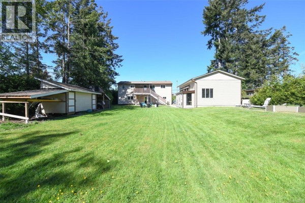 A wide view of a grassy backyard with a detached workshop and two houses, under a blue sky. - 1804 Little River Rd, Comox
