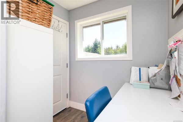 A small office space with pale grey walls, a white desk, a blue chair, and a window with trees outside. - 1804 Little River Rd, Comox