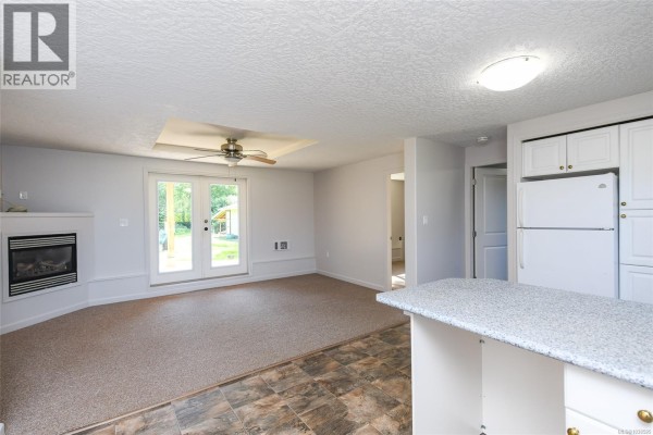 A living room area with a gas fireplace, beige carpet, white cabinets, and a kitchen island with granite countertop. - 1804 Little River Rd, Comox