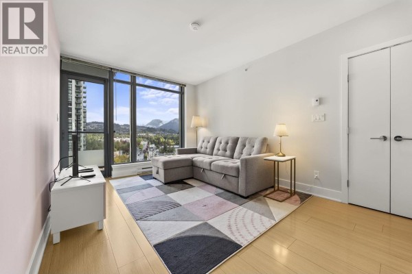 Living room with a grey sectional sofa, geometric rug, and mountain view through floor-to-ceiling windows. - 1806 1188 Pinetree Way, Coquitlam