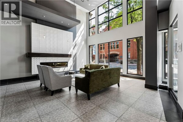 Building lobby with a modern fireplace, seating area, and large windows showing outdoor greenery. - 181 James Street N Unit# 312, Hamilton