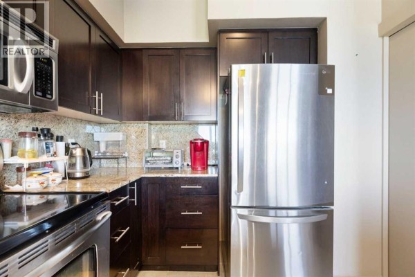 Kitchen with dark wood cabinets, granite countertops, and stainless steel refrigerator. - 1811, 8710 Horton Road Sw, Calgary