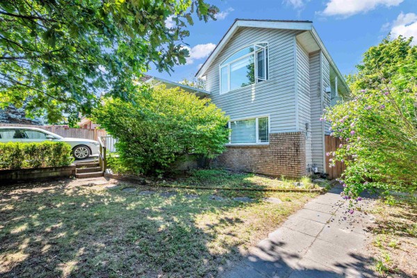 The side of a two-story house with light siding, brick base, and large windows is visible through trees. - 18136 Claytonwood Crescent, Surrey
