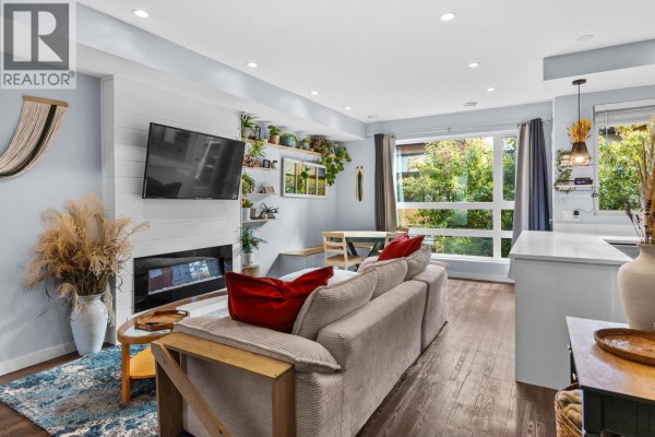 Living room with fireplace, TV, sofa, and dining area with large window. - 1818 Peak Point Court Unit# 30, West Kelowna