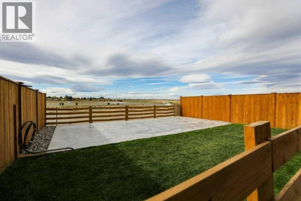 Fenced backyard with a concrete patio, grass area, and wooden ranch-style fencing with a distant view. - 182 Caribou Bend N, Lethbridge