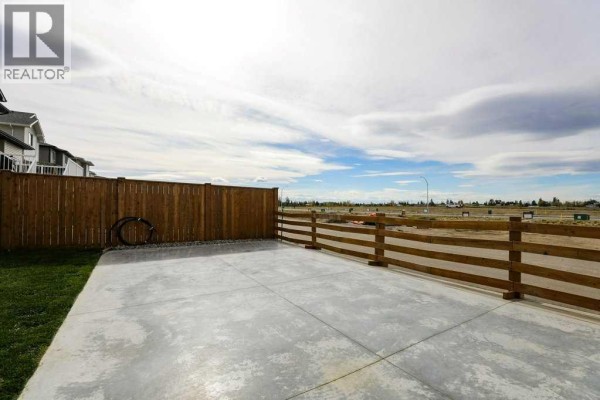 Corner view of a fenced backyard with a large concrete patio, grass, and wooden ranch-style fencing. - 182 Caribou Bend N, Lethbridge