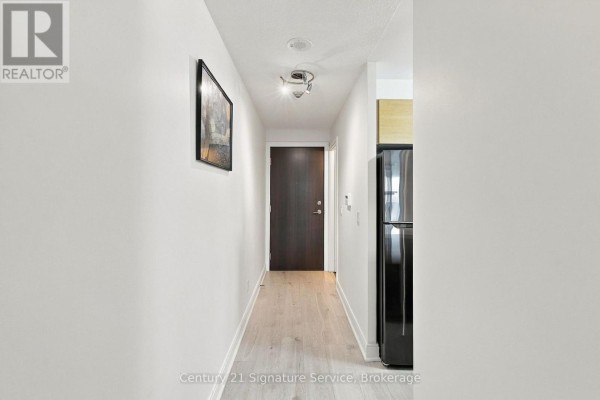 Interior view of an apartment hallway with white walls, light wood floors, and a black refrigerator. - 1822 - 135 Village Green Square, Toronto