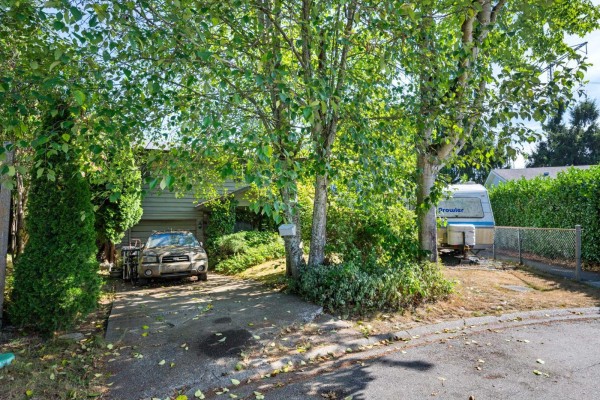 Front view of a modest single-story home with a driveway, car, trailer, and mature trees. - 18291 Bayard Place, Surrey