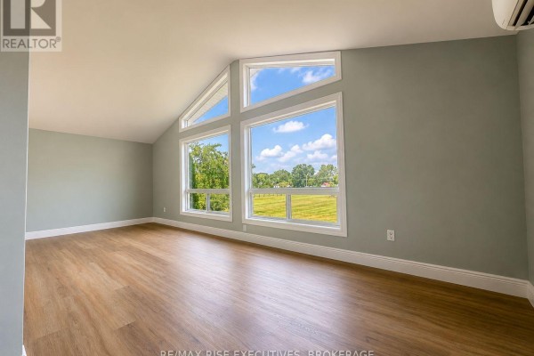 Empty room with light wood floors and large windows showing a rural landscape view. - 1848 County Road 9, Greater Napanee