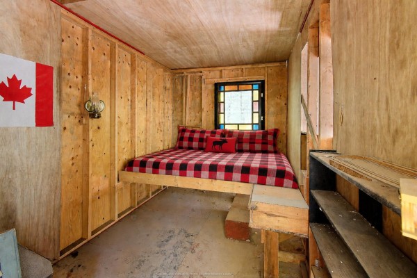 Interior view of a small cabin with unfinished wood walls, a raised bed, and a Canadian flag. - 1851 Ch. De La Rivière-noire, Waltham