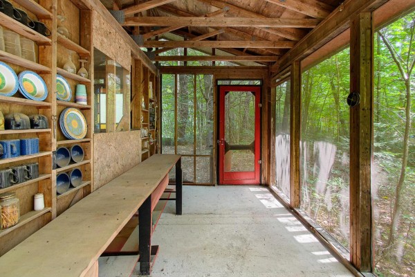 Interior screened porch area with wood-beamed ceiling, concrete floor, shelves filled with dishes, and a red door. - 1851 Ch. De La Rivière-noire, Waltham