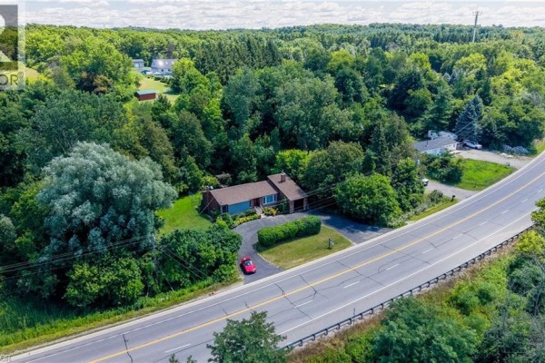 Aerial view showing the house, driveway, and surrounding wooded landscape beside a highway. - 1853 Highway 24 S, Cambridge