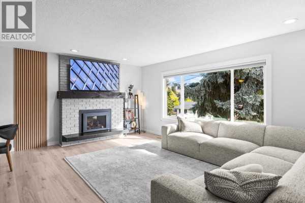 Living room with a large grey sectional sofa, fireplace, and big window with trees outside. - 1866 Large Avenue, Kelowna