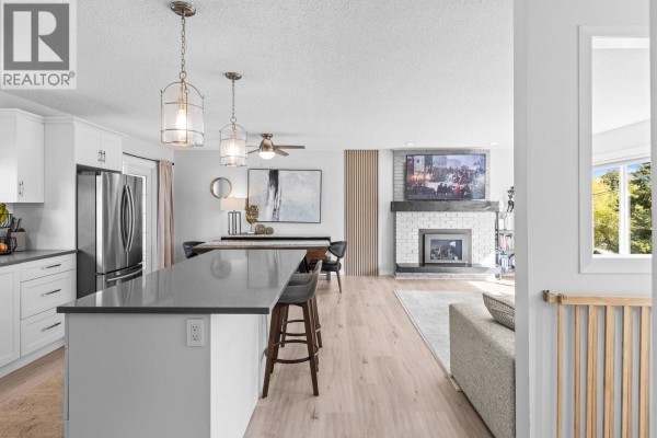Open-concept kitchen and living area with fireplace, dining table, and island with stools. - 1866 Large Avenue, Kelowna
