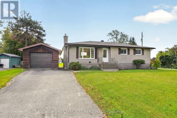 Front exterior of a light-colored brick bungalow with a paved driveway leading to a detached garage. - 18696 County Road 2 Road, South Glengarry
