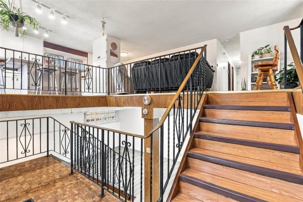 Interior view of a split-level staircase with wood steps, black railings, and a view of the kitchen and living area above. - 1884 Ross Avenue W, Winnipeg