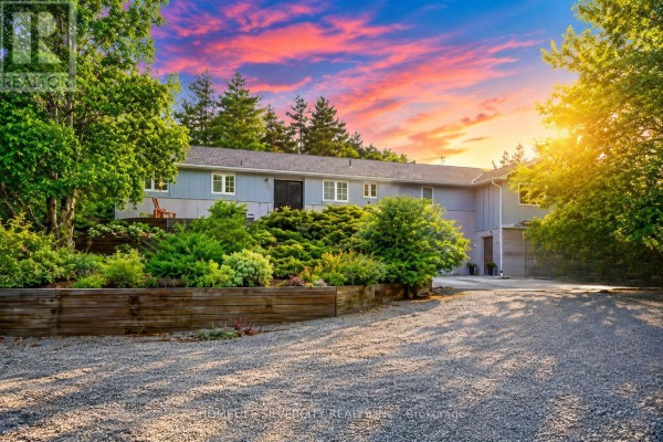 The grey exterior of the house is visible from the driveway with landscaping and a wooden retaining wall. - 18954 The Gore Road, Caledon