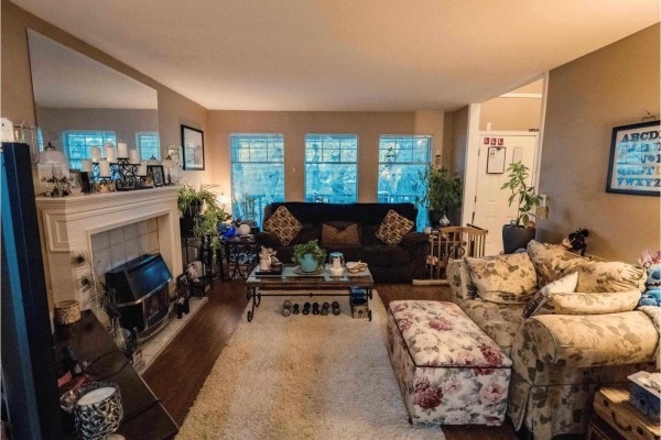 Living room with beige walls, large windows, and a fireplace, featuring floral sofas and decorative plants. - 18961 60b Avenue, Surrey