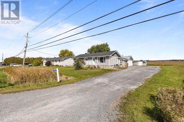 Driveway leading to a grey house with a large detached garage and rural background. - 1897 Champlain Road, Clarence Rockland