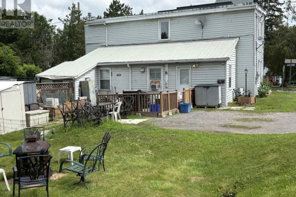 Rear exterior of a residential building with a metal roof extension, wooden deck, and grassy yard with outdoor furniture. - 19 & 21 Rock Lane E, Arnprior