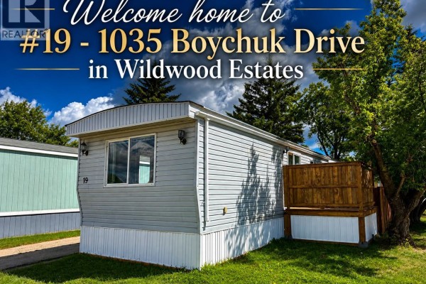 Side exterior view of a manufactured home with a wooden deck and trees. - 19 1035 Boychuk Drive, Saskatoon
