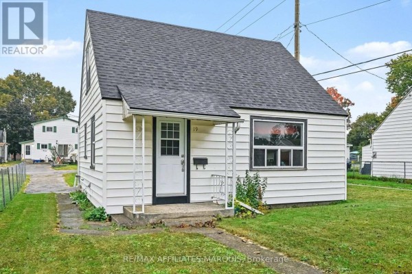 View of the front of a white sided bungalow with a covered porch and green lawn. - 19 Churchill Crescent, Smiths Falls