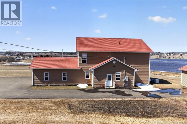 Rear view of the tan house and attached garage with a red roof, overlooking a body of water and distant shoreline. - 19 Leopold Goguen Road, Sainte Marie De Kent