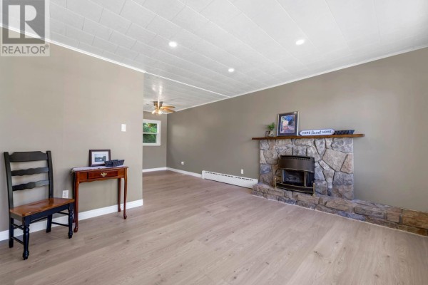 Living room with tan walls, light wood floors, a stone fireplace with a gas insert, and a wooden chair. - 19 Long Run Road, Petty Harbor   Maddox Cove