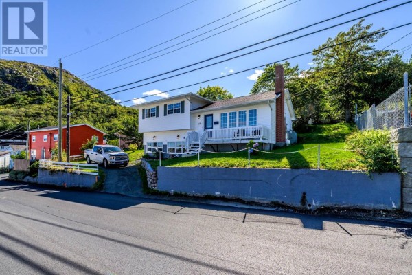 Side view of the white split-level house with a red neighbor building, driveway, and hilly landscape. - 19 Long Run Road, Petty Harbor   Maddox Cove