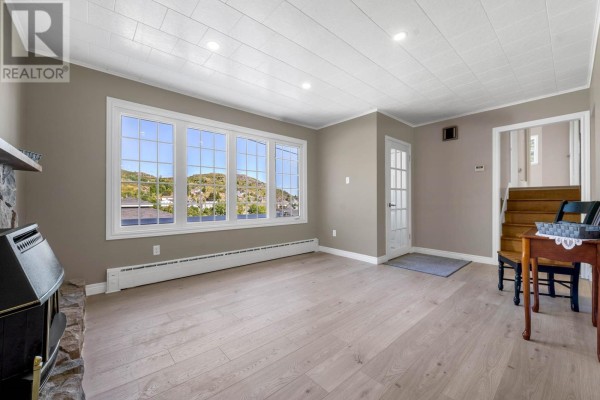 Living room with large bay windows facing hills, tan walls, light wood floors, and a glass-paned door to stairs. - 19 Long Run Road, Petty Harbor   Maddox Cove