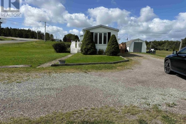 Front view of the white mobile home with a gravel driveway and two evergreen trees. - 19 Old Wharf Road, Havre Boucher