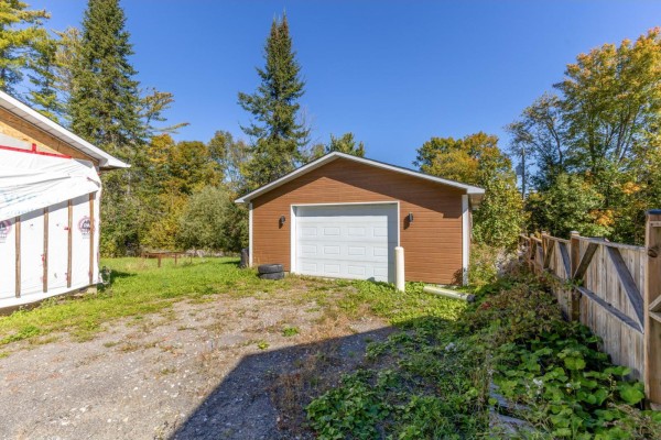 A detached brown garage with a white door and brown siding, situated next to a gravel driveway. - 19 Rue Du Centre, Val Des Monts