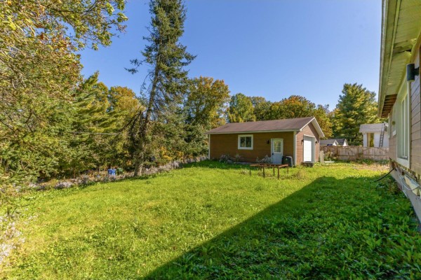 View of a backyard with a sloped green lawn, trees, and a detached garage with a white door. - 19 Rue Du Centre, Val Des Monts
