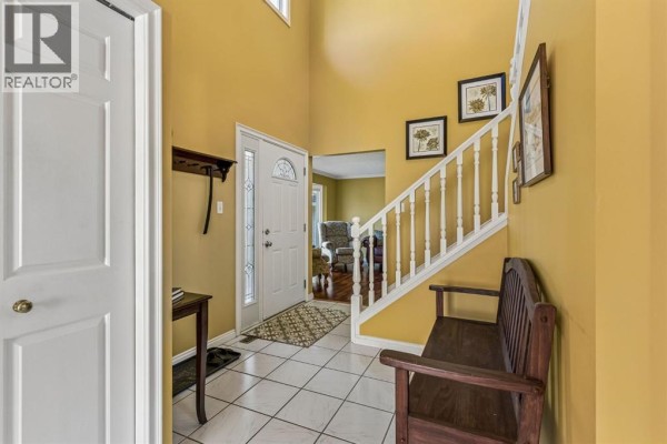 Entrance foyer with yellow walls, white tiled floor, white door, and wooden bench by the stairs. - 19 Woodbrook Road Sw, Calgary
