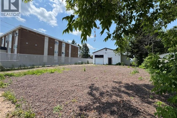 Empty lot with gravel surface, a detached garage, and a brick apartment building behind fence. - 190 Spruce Street, Moncton